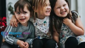 Three children share a group hug, smiling and laughing on a doorstep on a sunny day. The color photo shows them dressed casually.