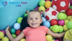 A blonde girl plays in a therapy playroom, sitting in a ball pit and smiling happily at the camera during an autism intervention session.