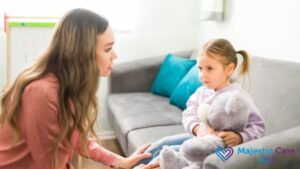 ABA therapy session, a female therapist gently soothes a young girl sitting on a sofa while she hugs a teddy bear.