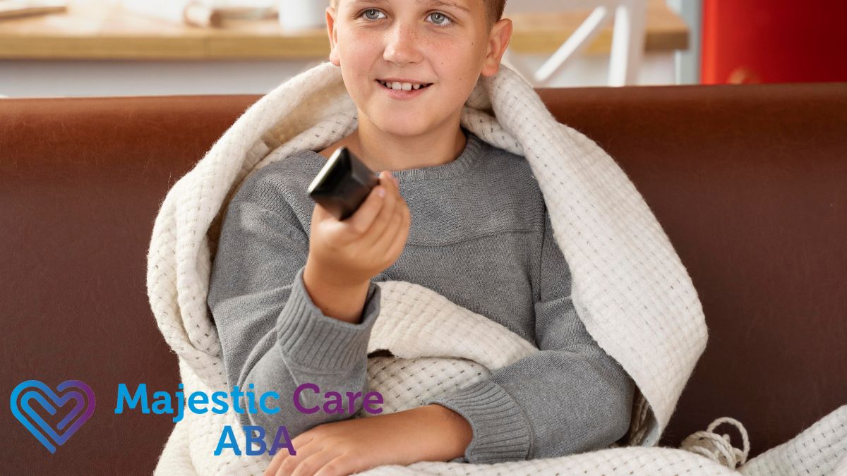A young boy with autism sits on a couch holding a remote, wrapped in a heavy cream-colored weighted blanket.