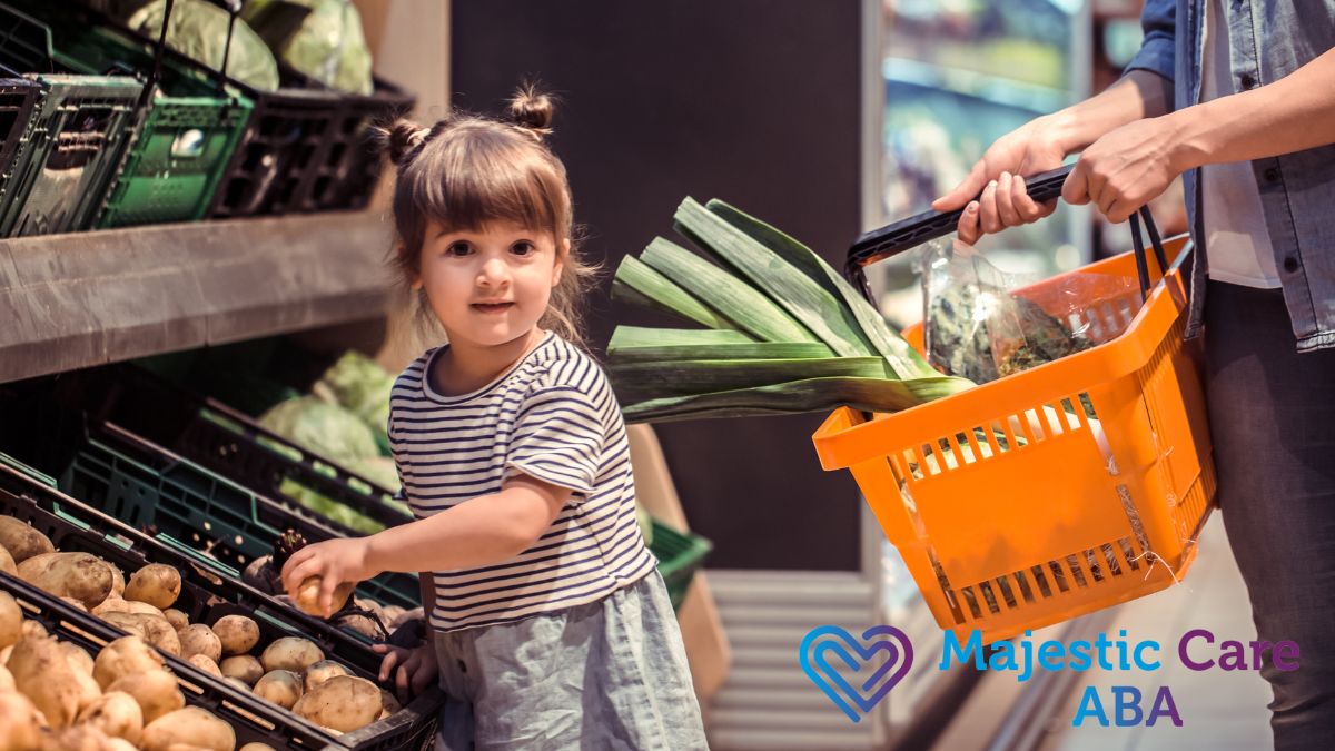 A young girl practices autism life skills by selecting vegetables during grocery shopping while an adult holds out a basket for her.