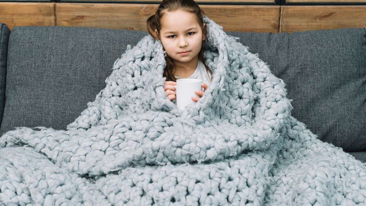 A young girl with autism wrapped in a weighted blanket holds a mug and stares intensely at the camera.