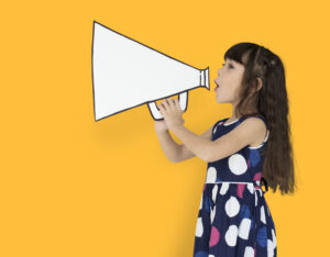 A young girl against a yellow background holding an illustration of a large megaphone, demonstrating verbal behavior in ABA.