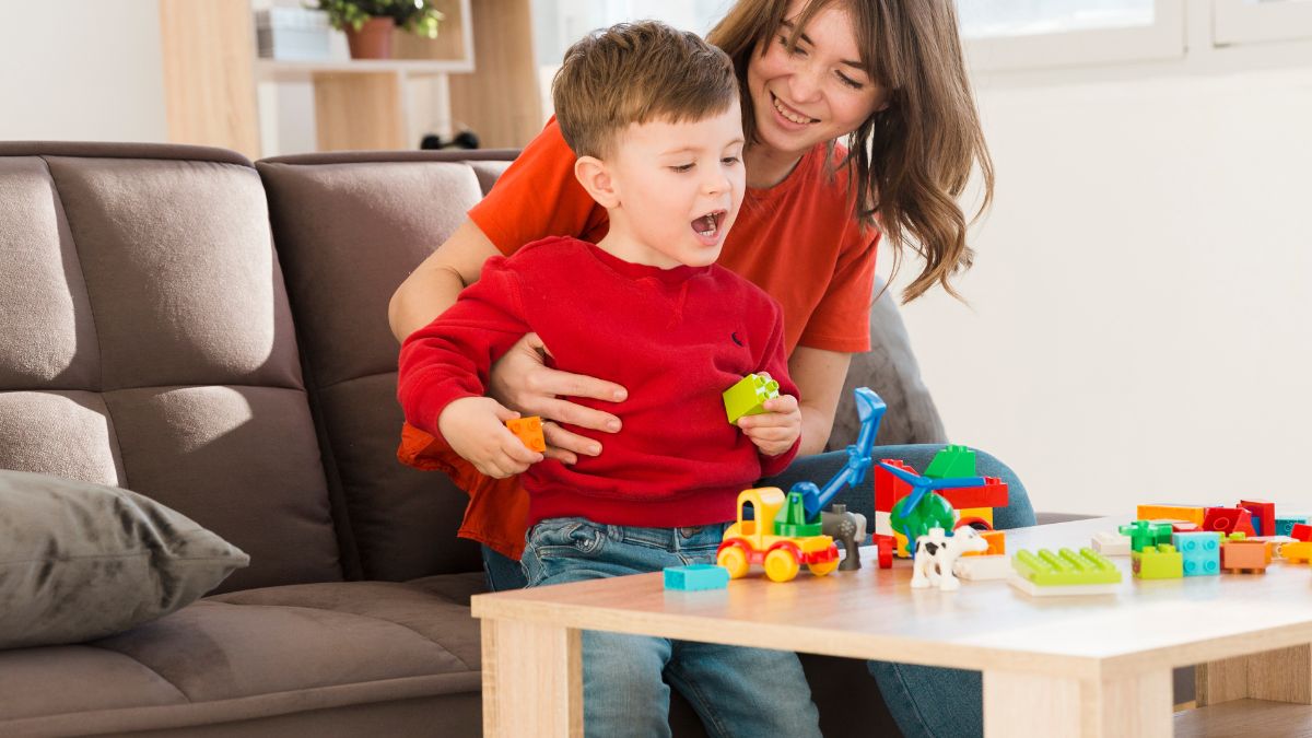 A parent or therapist guides a young child playing with puzzle pieces and blocks on a table in a brightly lit living room.