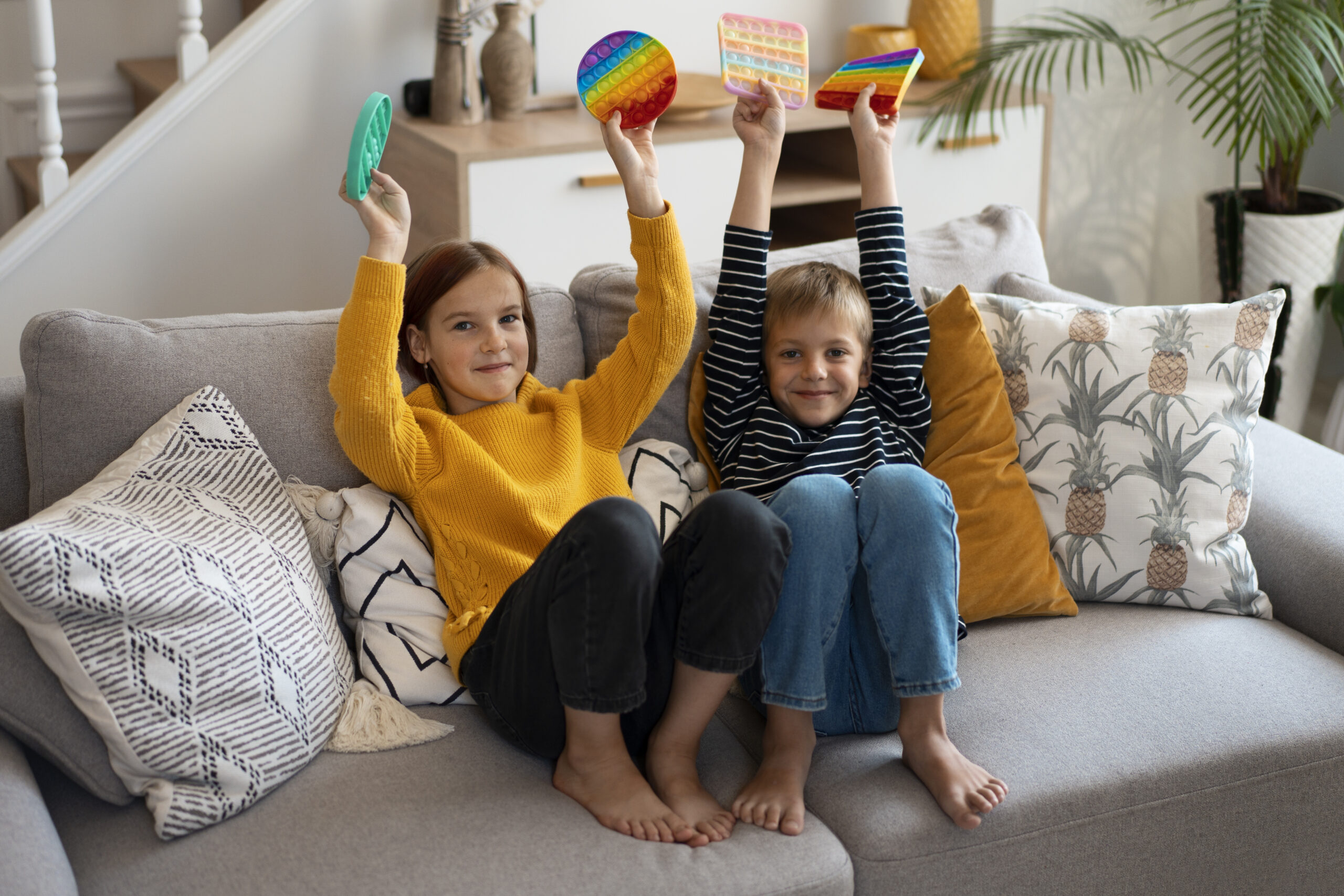 Two children sit on a comfy gray sofa during in-home ABA therapy, raising their hands while holding fidget toys.